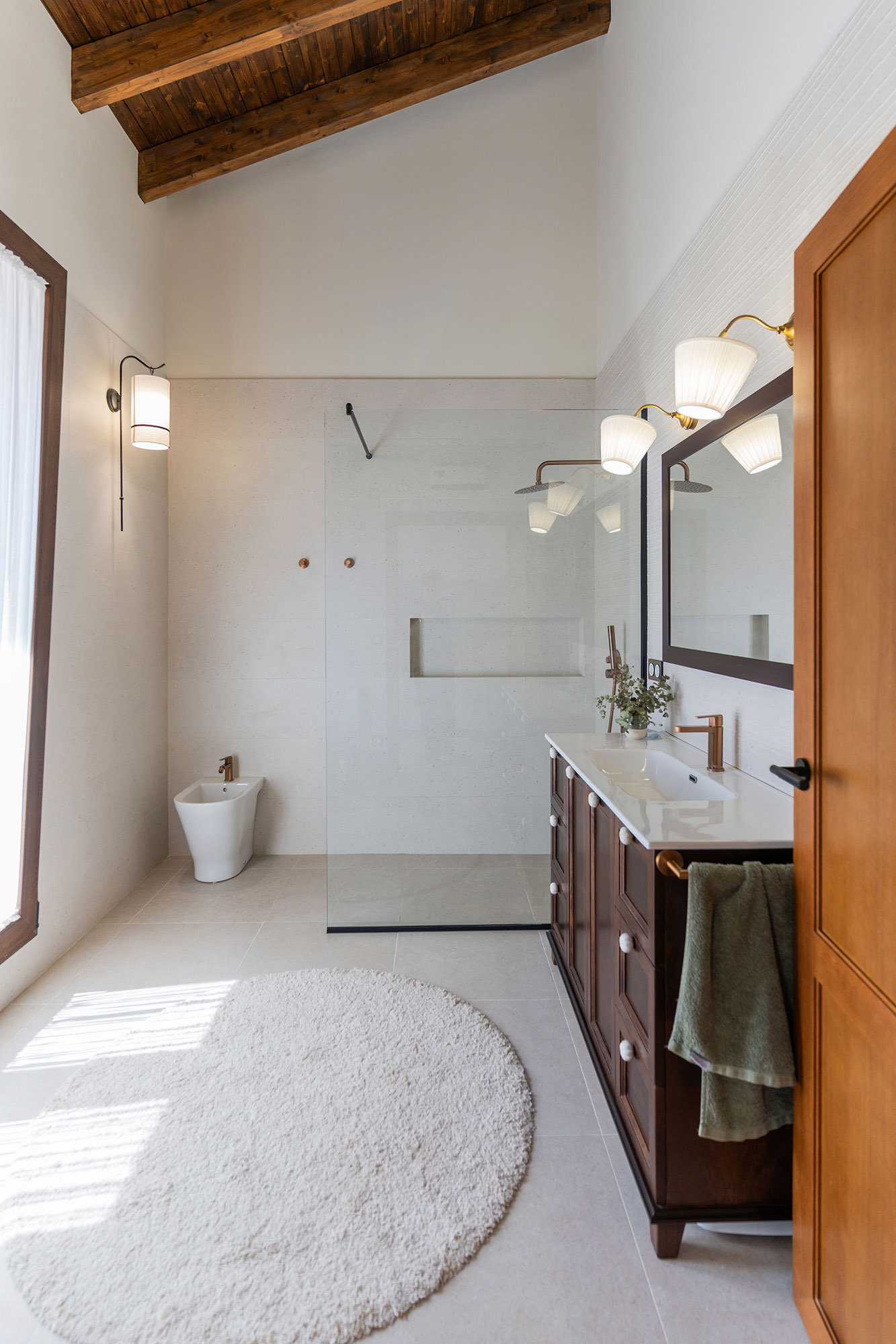 Baño reformado de Casa Sofia con ducha a ras de suelo, revestimientos claros, mueble de madera y techo con vigas vistas.